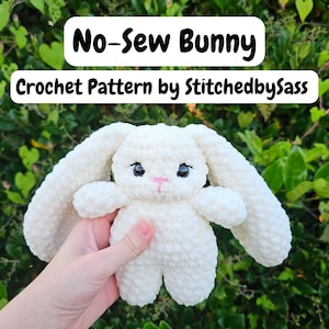 Peut inclure: Un lapin en peluche blanc fait de fil doux avec de longues oreilles, des yeux en plastique noir et un nez brodé rose. Le texte au-dessus indique No-Sew Bunny Crochet Pattern by StitchedbySass. Le lapin a une tête et un corps arrondis.