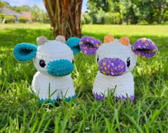 Puede incluir: Dos peluches de vaca tejidos a mano con cuerpos blancos y ojos de seguridad negros. Una vaca tiene orejas, hocico y pezuñas de color verde azulado con cuernos color canela. La otra tiene orejas, hocico y pezuñas moradas moteadas con cuernos naranjas.