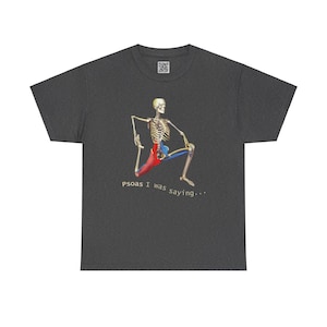 Puede incluir: Una camiseta gris oscuro jaspeada con un esqueleto haciendo una pose de yoga. El músculo psoas del esqueleto está resaltado en rojo. El texto "Psoas I was saying..." está impreso debajo del esqueleto.