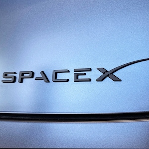 May include: A black and white SpaceX logo on a blue background. The logo features the word "SpaceX" in a bold, sans-serif font and a stylized rocket ship.