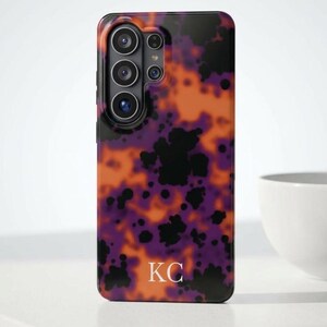 Op de afbeelding: Een telefoonhoesje met een levendig abstract ontwerp in oranje, paars en zwart. De hoes heeft een cameramodule en de initialen "KC" onderaan. De hoes ligt op een wit oppervlak.
