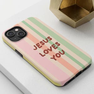 May include: A smartphone case featuring vertical stripes in pastel shades of green, peach, and pink. The text "JESUS LOVES YOU" is printed in a bold, dark red font across the center of the case. The case has a black camera cutout and black edges.