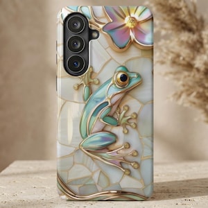 May include: A phone case featuring a detailed, three-dimensional design of a frog and flower. The frog is light blue and green with gold accents, set against a white and gold mosaic background. A flower with iridescent petals is also featured.