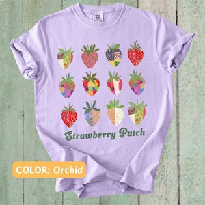 May include: A lavender t-shirt with a graphic of a strawberry patch. The strawberries are made of colorful patchwork fabric. The text "Strawberry Patch" is printed in green below the strawberries. The text "COLOR: Orchid" is printed in yellow at the bottom of the shirt.