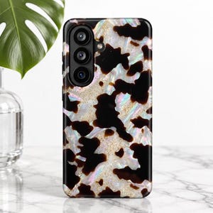 May include: A smartphone case featuring a black and iridescent white cow print pattern with gold glitter accents. The case has a cutout for a triple-lens camera system on the upper left side and a smooth, glossy finish throughout.