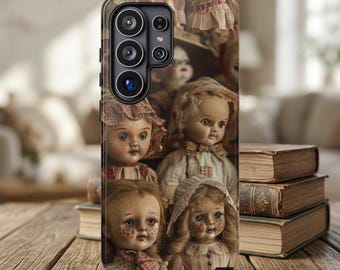 May include: A smartphone case featuring a collage of vintage-style dolls with painted facial features, wearing bonnets and textured clothing. The design displays multiple doll faces with detailed eyes and rosy cheeks in a layered, artistic arrangement.