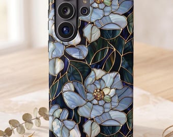 Op de afbeelding: Een rechthoekig smartphonehoesje met een patroon geïnspireerd op glas-in-lood, bestaande uit lichtblauwe bloemen met witte harten en donkergroene bladeren, omlijnd met dunne gouden lijnen die de gehele achterkant van de hoes bedekken.