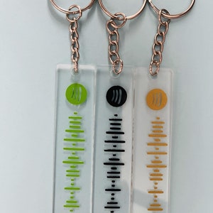 May include: Three keychains with clear rectangular acrylic charms featuring a stylized music note and a vertical barcode pattern. The charms are green, black, and gold.