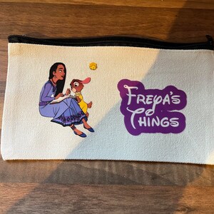 May include: A white canvas zippered pouch with a purple design that says "Freya's Things". The pouch features an illustration of a woman with long dark hair in a blue dress sitting with a rabbit.