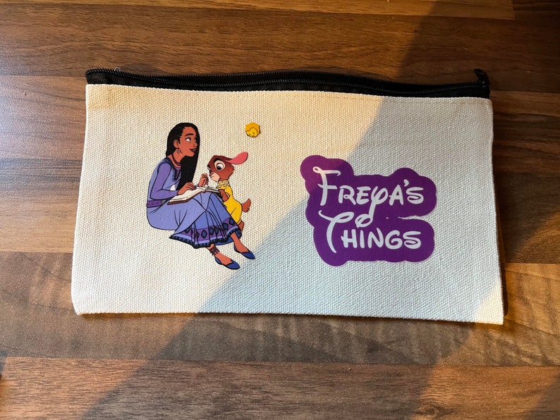 May include: A white canvas zippered pouch with a purple design that says "Freya's Things". The pouch features an illustration of a woman with long dark hair in a blue dress sitting with a rabbit.