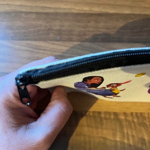 May include: A white canvas zippered pouch with a colorful cartoon image of a woman and a rabbit. The pouch is partially open, showing the black zipper.