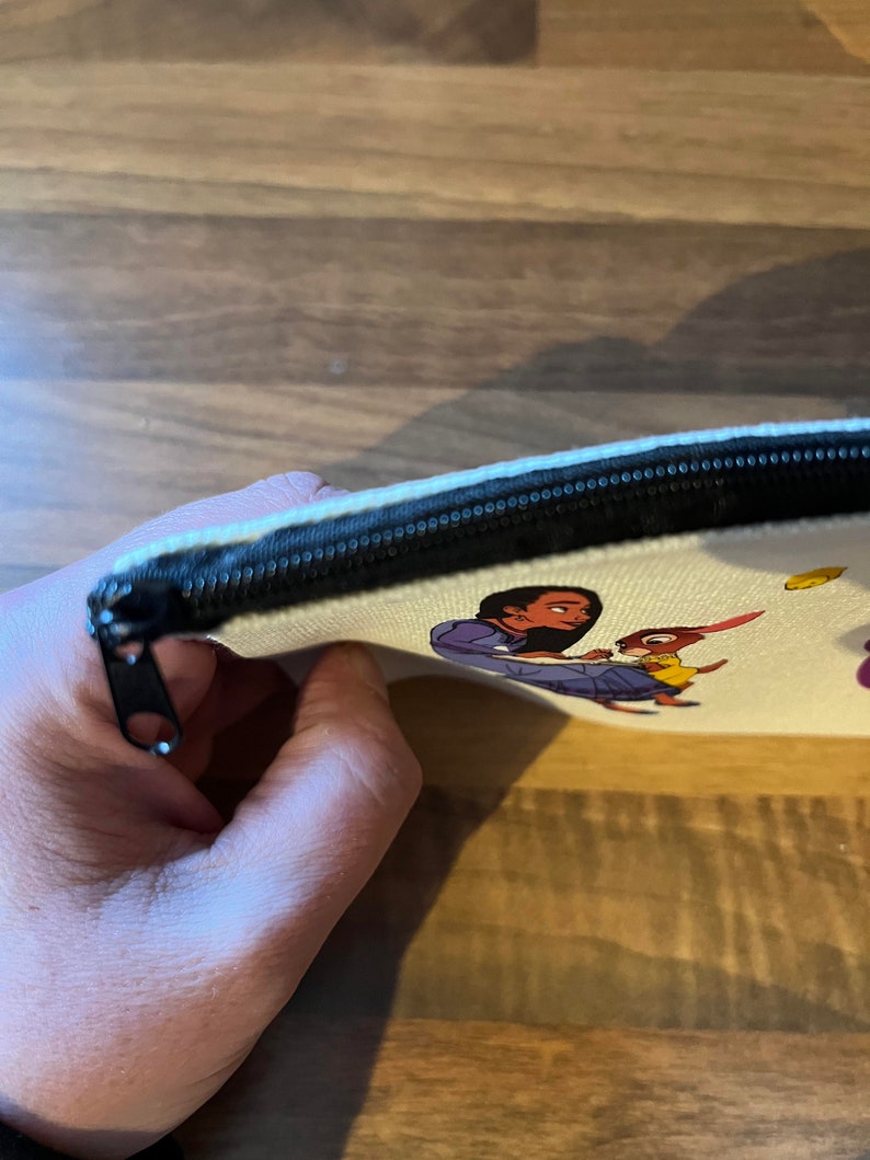 May include: A white canvas zippered pouch with a colorful cartoon image of a woman and a rabbit. The pouch is partially open, showing the black zipper.