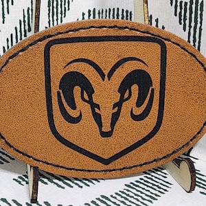 May include: A brown, oval-shaped patch featuring a black ram's head logo within a black border. The patch is made of a textured material and is stitched with a dark thread. The background is a white fabric with a green geometric pattern.