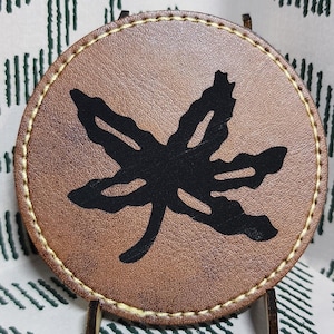 May include: A round, brown leather coaster with a black leaf design. The coaster has a stitched edge and is displayed on a small metal stand. The background is a white fabric with a green geometric pattern.