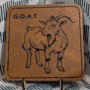 May include: Square, brown leather coaster with a black illustration of a goat and the letters "G.O.A.T." The coaster is displayed on a small wooden stand against a patterned background.