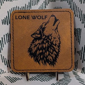 May include: A square, tan leather coaster with the words "LONE WOLF" above a howling wolf illustration. The wolf is black and detailed. The coaster is propped up on a small wooden stand against a patterned background.