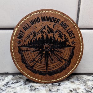 May include: A circular brown leather patch with light-colored stitching around the edge. The center features a black laser-engraved design of mountains, a forest, and a compass over a wood grain pattern with the text NOT ALL WHO WANDER ARE LOST.