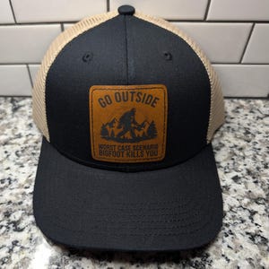 May include: A black and tan trucker hat featuring a brown leather-like patch on the front. The patch displays a graphic of a mountain landscape with a Bigfoot silhouette and the text: GO OUTSIDE WORST CASE SCENARIO BIGFOOT KILLS YOU.