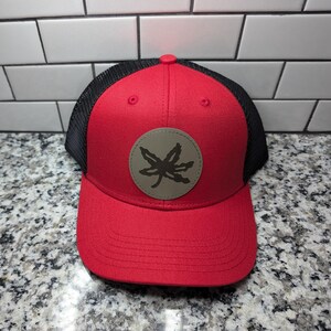 May include: A red baseball cap with a black mesh back and a circular tan patch on the front. The patch features a dark brown graphic of a jagged leaf. The cap has a structured front panel, a curved brim, and a button at the top center.