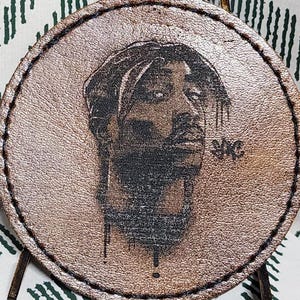 May include: A round, light brown leather coaster featuring a portrait of Tupac Shakur. The portrait is a detailed, black and white illustration. The coaster has a stitched border and is set against a patterned fabric background.