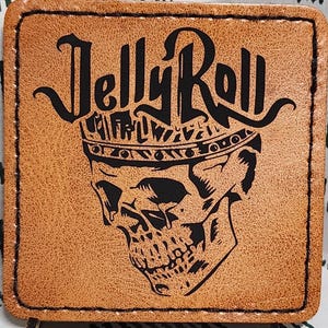 May include: A square, tan leather patch with the words "Jelly Roll" above a skull wearing a crown. The patch is propped up on a patterned fabric with a white background and dark green diagonal lines.