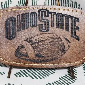 May include: A rectangular, brown leather patch with the words "OHIO STATE" and a football graphic. The patch is stitched around the edges and is displayed against a white and green patterned background.