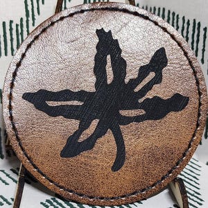 May include: A round, brown leather coaster with a black, stylized leaf design. The coaster is displayed on a small wooden stand against a white fabric with a green geometric pattern. The coaster is approximately 4 inches in diameter.