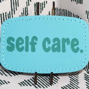 May include: A light blue, rectangular object with rounded corners, stitched edges, and the words "self care." in green. The object is set against a white fabric with a black zigzag pattern.