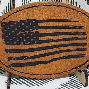 May include: Oval-shaped leather patch with a distressed American flag design. The flag features black stars and stripes on a tan background, with a stitched border. The patch is displayed on a wooden stand.