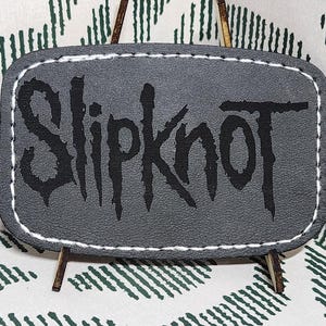May include: A gray leather patch with the band name "Slipknot" in black, stitched with white thread. The patch is rectangular with rounded corners. It is displayed on a small wooden easel against a patterned background.