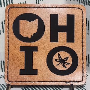 May include: A square, light brown leather coaster with the word "OHIO" in black block letters and a black outline of the state. The coaster is supported by small metal legs and sits on a patterned fabric.