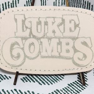 May include: A cream-colored rectangular tag with the name "LUKE COMBS" in a stylized font. The tag is displayed against a white fabric with a green geometric pattern. The tag is held up by a small stand.