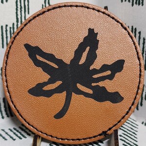 May include: A round, brown leather coaster with a black leaf design. The coaster is displayed on a small wooden stand against a patterned white and green background. The coaster is approximately 4 inches in diameter.