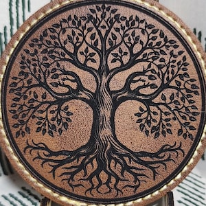May include: A round, brown coaster with a detailed black tree of life design. The coaster has a light brown border with a stitched pattern. It is displayed on a small wooden stand, set against a white and green patterned background.
