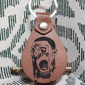 May include: A brown leather keychain with a silver ring. The keychain features a black graphic of a person's face with hands covering the eyes and the word "BOO" visible. The keychain is on a wooden stand.