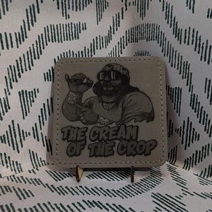 May include: A square, light gray coaster featuring a black and white illustration of a man wearing sunglasses and a hat, flexing his arm. The text "THE CREAM OF THE CROP" is below the illustration. The coaster is displayed on a small stand.