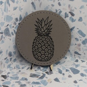 May include: A circular gray coaster featuring a black laser-engraved pineapple illustration in the center. The coaster has visible stitching along the outer edge and is supported by a small stand, displaying a detailed graphic of the fruit.