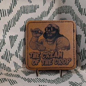 May include: Square, brown leather coaster with a graphic of a man wearing sunglasses and a camouflage hat. The text "THE CREAM OF THE CROP" is below the graphic. The coaster is on a metal stand.