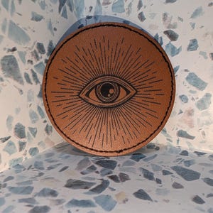 May include: A circular tan leather patch featuring a black engraved design of an eye surrounded by radiating sunburst lines. The patch has a visible black stitched border around the entire circumference, creating a distinct contrast against the tan.