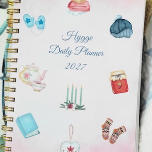 May include: A spiral-bound daily planner with a light pink cover featuring the text Hygge Daily Planner 2027. The cover displays watercolor illustrations of a folded blanket, blue mittens, a blue hat, a teapot, a cup, four candles, a jar of jam, a book, a heart ornament, and patterned socks.
