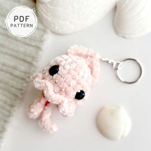 May include: A handmade pink crochet squid keychain with black eyes and a silver keyring. The keychain is made of soft, plush yarn. A white circle with the text "PDF PATTERN" is in the background.