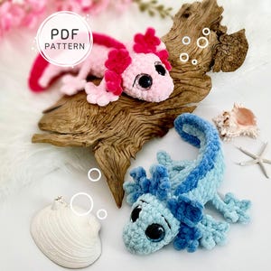 Puede incluir: Dos peluches de axolote tejidos a ganchillo, uno rosa claro con branquias rosa oscuro y otro azul claro con branquias azul oscuro, con grandes ojos negros. Una etiqueta blanca circular dice PDF PATTERN. Los peluches están sobre madera flotante.