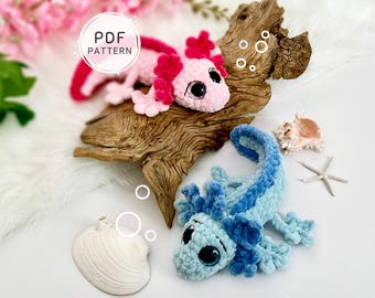May include: Two crocheted axolotl plushies, one light pink with dark pink gills and one light blue with dark blue gills, featuring large black eyes. A circular white label reads PDF PATTERN. The plushies are arranged with a piece of driftwood.