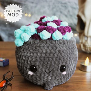 May include: A grey, plush cauldron toy with a smiling face and large black eyes. The cauldron is topped with teal and purple plush balls. A small black spider with an orange gem is in the foreground. The text "PATTERN MOD" is in a white starburst.