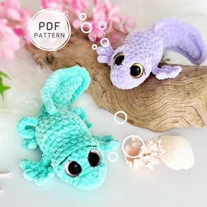 May include: Two crocheted plush axolotl toys in light mint green and soft lavender colors. Each creature features large, dark, glossy eyes and textured, fuzzy yarn bodies. A circular graphic with the text PDF PATTERN is visible.