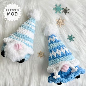 May include: Two handmade crochet gnomes with blue and white striped hats and white beards. Each gnome has a pink nose and black feet. The gnomes are on a white, fluffy surface with decorative snowflakes. The text "PATTERN MOD" is on a white star.