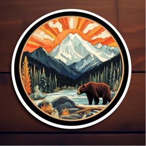 May include: A round sticker with a colorful illustration of a mountain range with a bear in the foreground. The mountains are blue and white with a sunset behind them. The bear is brown and is standing in a river.