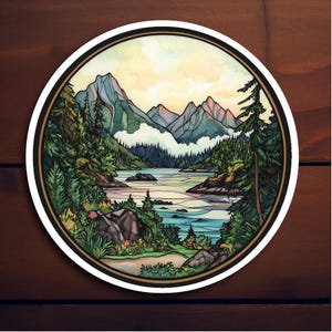 May include: Round sticker featuring a watercolor landscape of mountains, a lake, and evergreen trees. The design has a black border with a white outline. The sky is a gradient of yellow and orange. The sticker is on a wooden surface.