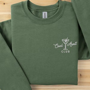 May include: An olive green sweatshirt with the words "Cool Aunt Club" and a cocktail glass graphic in white. The sweatshirt is folded on a light brown wooden surface.
