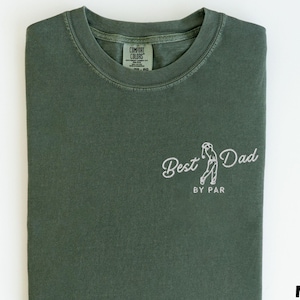 May include: Moss green t-shirt with the words "Best Dad" and a golf-themed graphic embroidered in white. The phrase "BY PAR" is below the graphic. The shirt is folded neatly on a white surface.